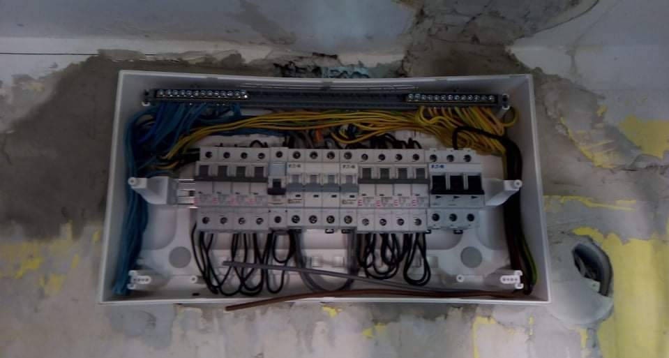 Rozdzielnica elektryczna w białej obudowie, z widocznymi przewodami i bezpiecznikami, zamontowana na surowej ścianie w trakcie prac instalacyjnych.