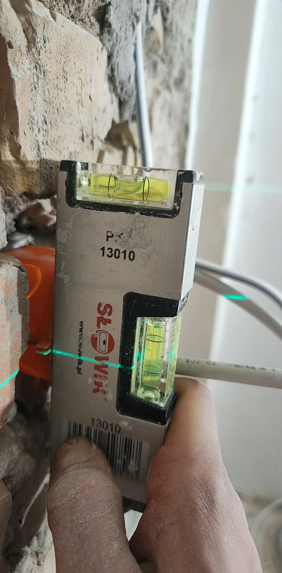 Poziomica laserowa w dłoni na tle surowej ściany z cegły i kabli. Precyzyjne wyznaczanie poziomu podczas prac instalacyjnych.
