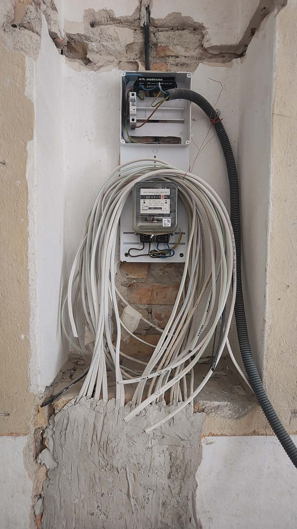 Licznik elektryczny z plątaniną białych kabli na tle surowej ściany w trakcie prac instalacyjnych. Widoczna skrzynka rozdzielcza i peszel.