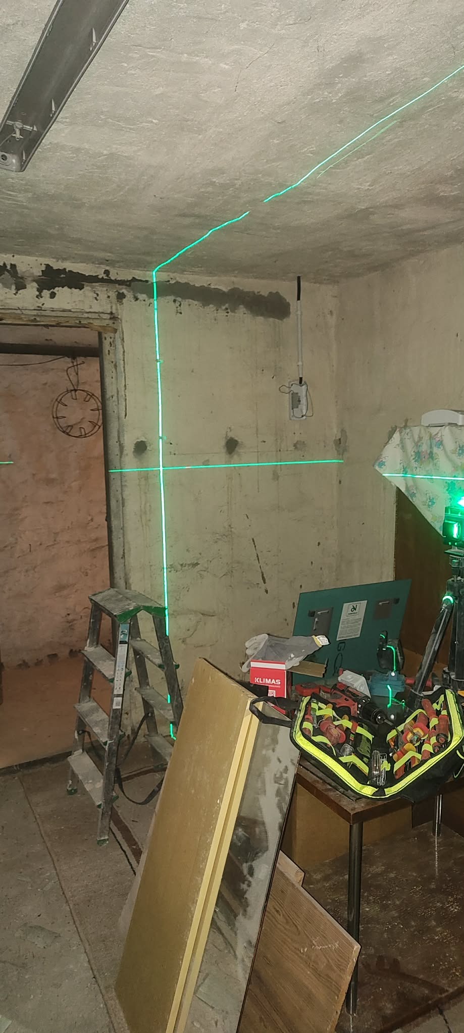 Laser krzyżowy wyznacza poziom na ścianach w trakcie remontu. Widoczna drabina, narzędzia i torba narzędziowa. Surowy stan ścian przed wykończeniem.