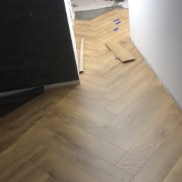 K2. - Ułożone panele podłogowe w jodełkę, widok z góry na fragment podłogi i buta. W tle widoczne materiały budowlane i okno.