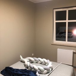 K2. - Pokój po malowaniu ścian na beżowo, widoczne okno z grzejnikiem, łóżko z ozdobnym warkoczem i pościelą. Ściany świeżo pomalowane, brak śladów niedociągnięć.