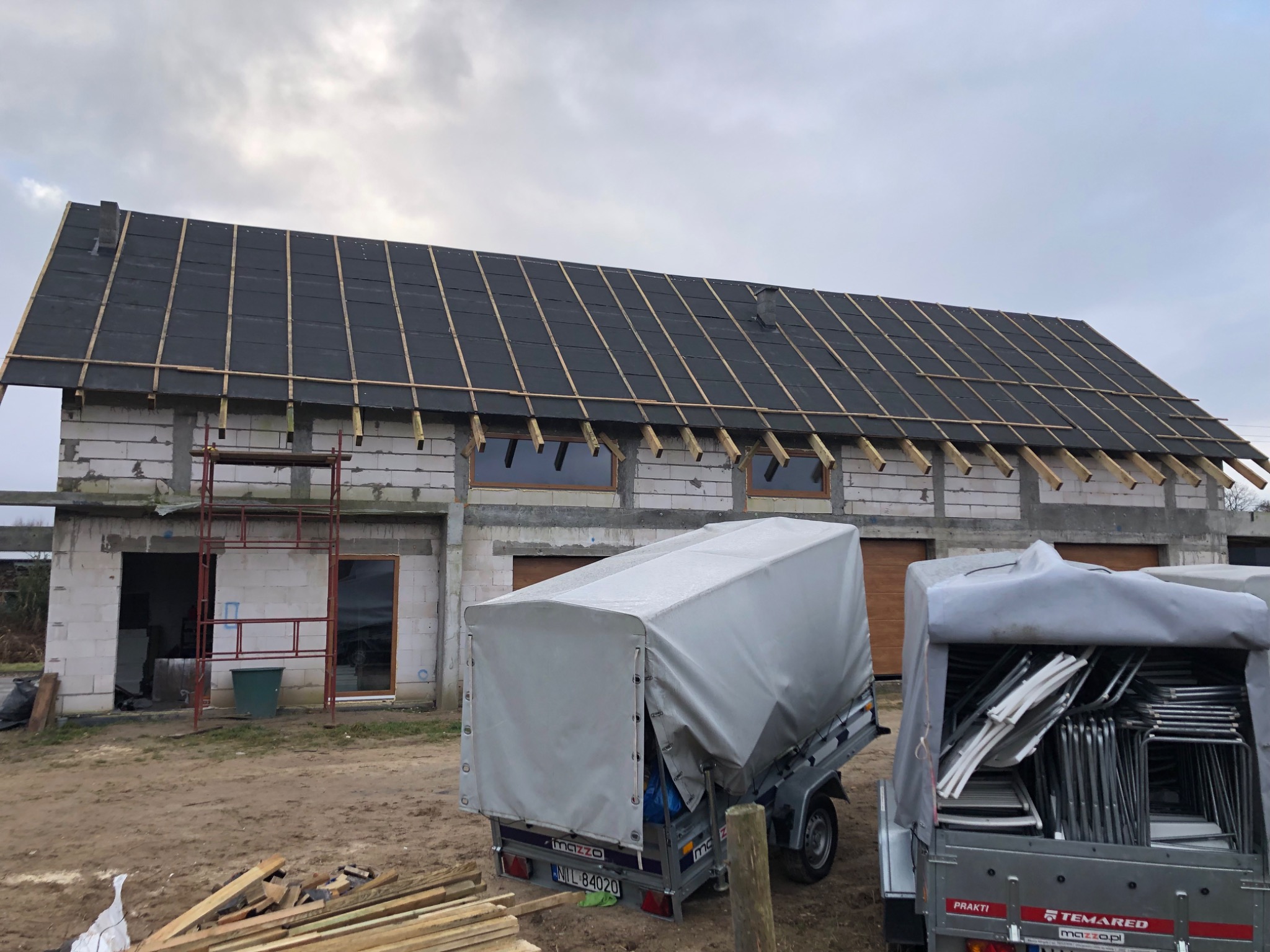 Budynek w trakcie budowy dachu, widoczne krokwie i papa. Na pierwszym planie przyczepki z materiałami budowlanymi. Surowy stan, cegły silikatowe.
