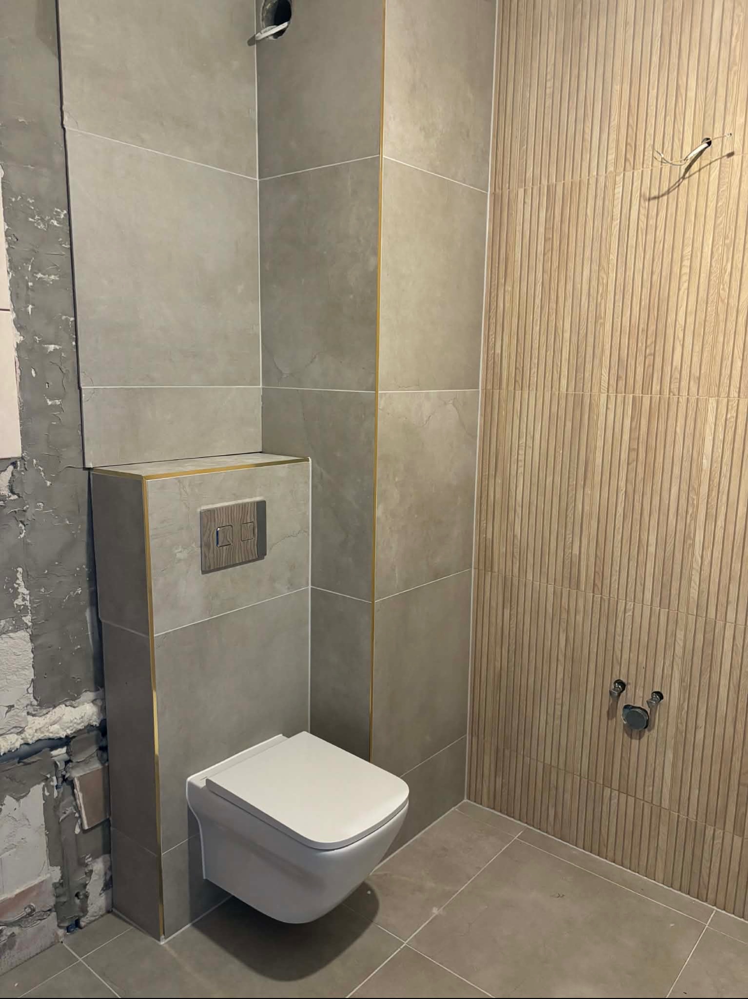 Nowoczesna toaleta podwieszana z szarą płytką imitującą kamień i drewnianą ścianą z lamelami. Widoczne niedokończone prace na ścianie po lewej stronie.