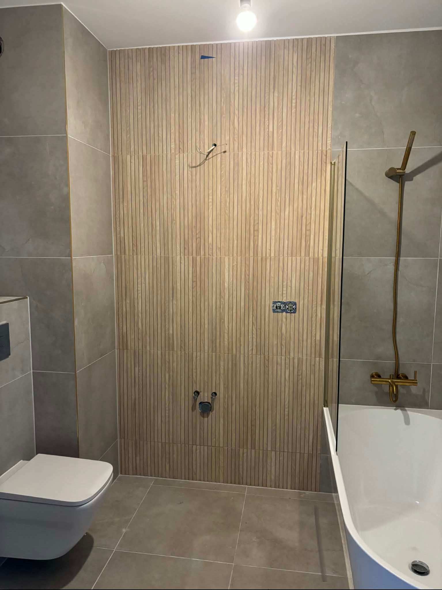 Nowoczesna łazienka z drewnopodobnymi panelami ściennymi, szarą glazurą i złotą armaturą. Minimalistyczny design, widoczna wanna i toaleta podwieszana.