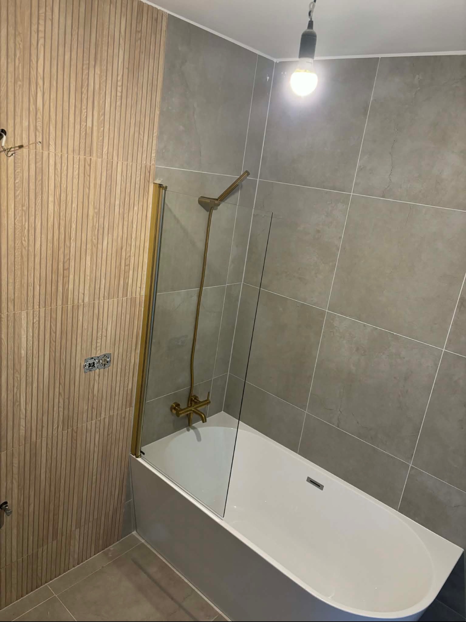 Wykończona łazienka z wanną, szklanym parawanem i złotą armaturą. Ściany wyłożone płytkami w odcieniach szarości i drewnopodobnymi panelami. Widoczne gniazdka elektryczne.