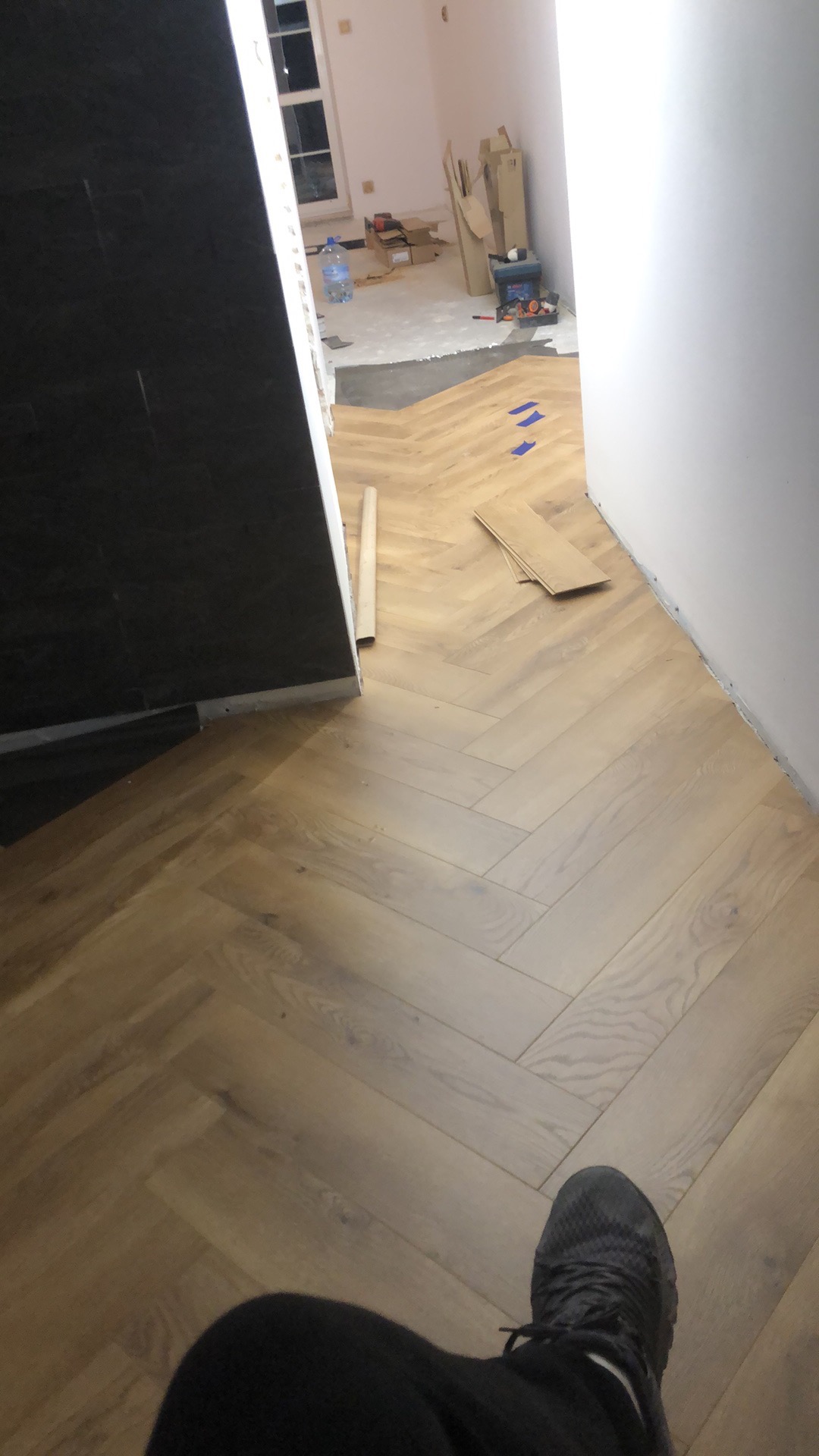 Ułożone panele podłogowe w jodełkę, widok z góry na fragment podłogi i buta. W tle widoczne materiały budowlane i okno.