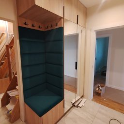 Malwa meble - Funkcjonalna zabudowa przedpokoju z siedziskiem obitym butelkową zielenią, wbudowanym lustrem i szafkami z jasnego drewna. Widoczne schody i fragment podłogi.