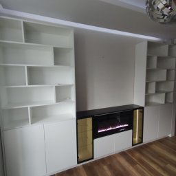 Malwa meble - Biała zabudowa meblowa z półkami i szafkami, wkomponowana we wnękę ściany, z czarnym blatem i elektrycznym kominkiem. Nowoczesny design, drewniana podłoga.