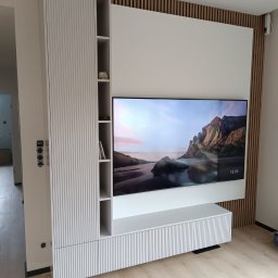 Malwa meble - Biała zabudowa RTV z lamelami i szafką wiszącą, zintegrowana z drewnianą ścianką działową. Nowoczesny design, minimalistyczny styl, widok z perspektywy podłogi.