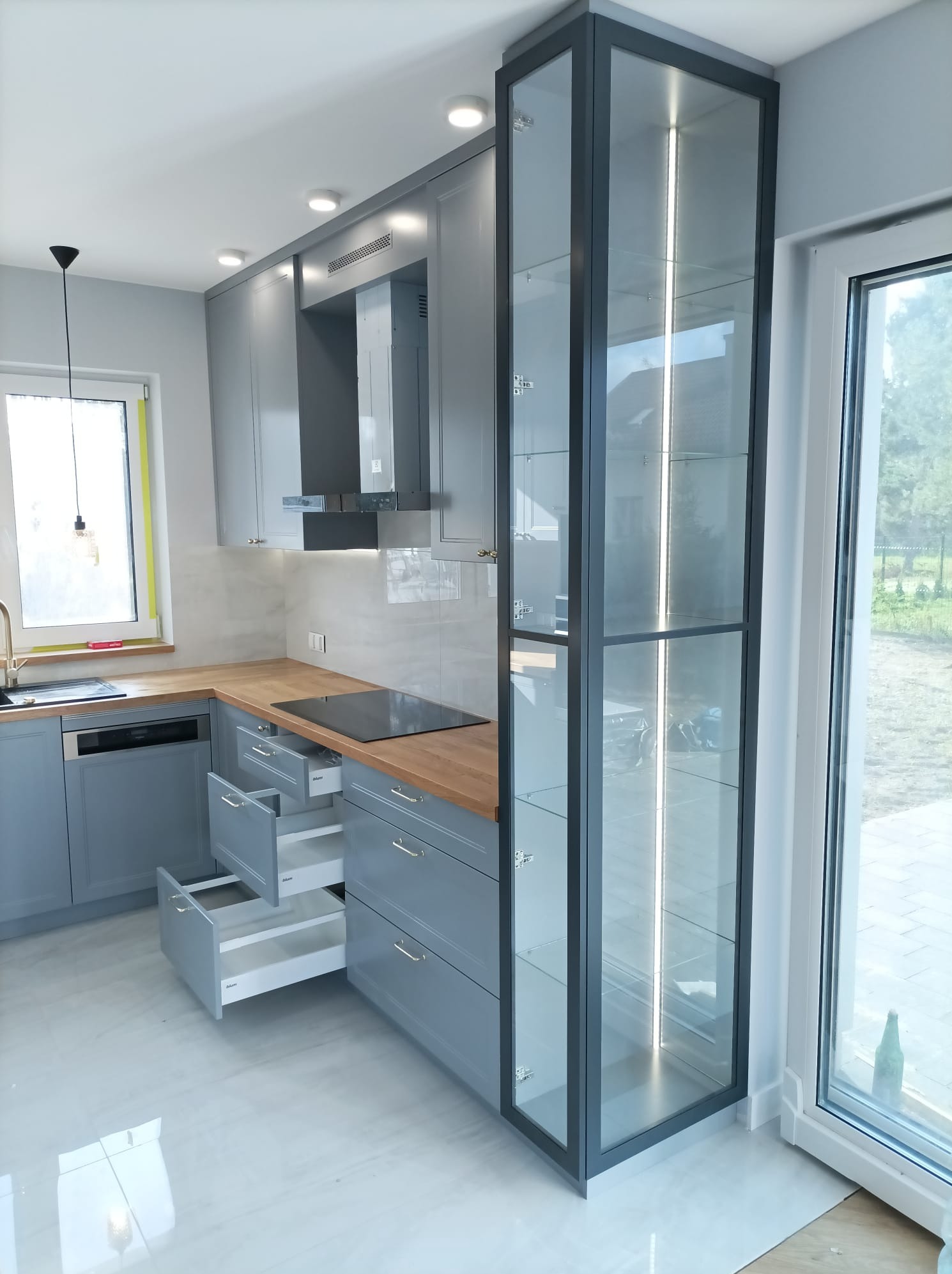 Szara kuchnia z drewnianym blatem, otwartymi szufladami i wysoką, przeszkloną witryną z oświetleniem LED, widoczna z perspektywy narożnej.