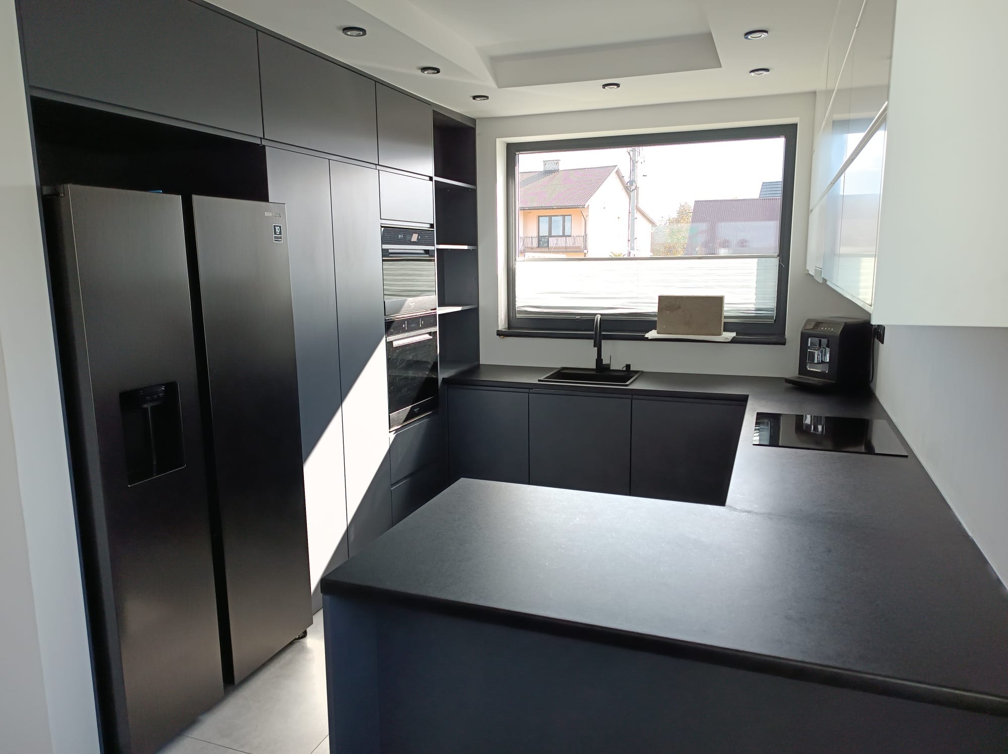 Nowoczesna, czarna kuchnia z zabudową po sufit i dużym oknem. Minimalistyczny design, ciemne blaty i sprzęty AGD tworzą elegancką całość. Widok na dom z zewnątrz.