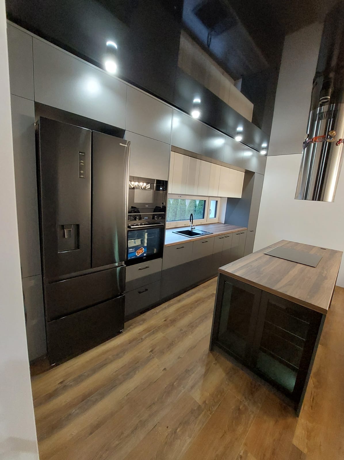 Nowoczesna kuchnia z szarymi szafkami, czarną lodówką side-by-side i wyspą z drewnianym blatem. Błyszczący sufit z oświetleniem punktowym.