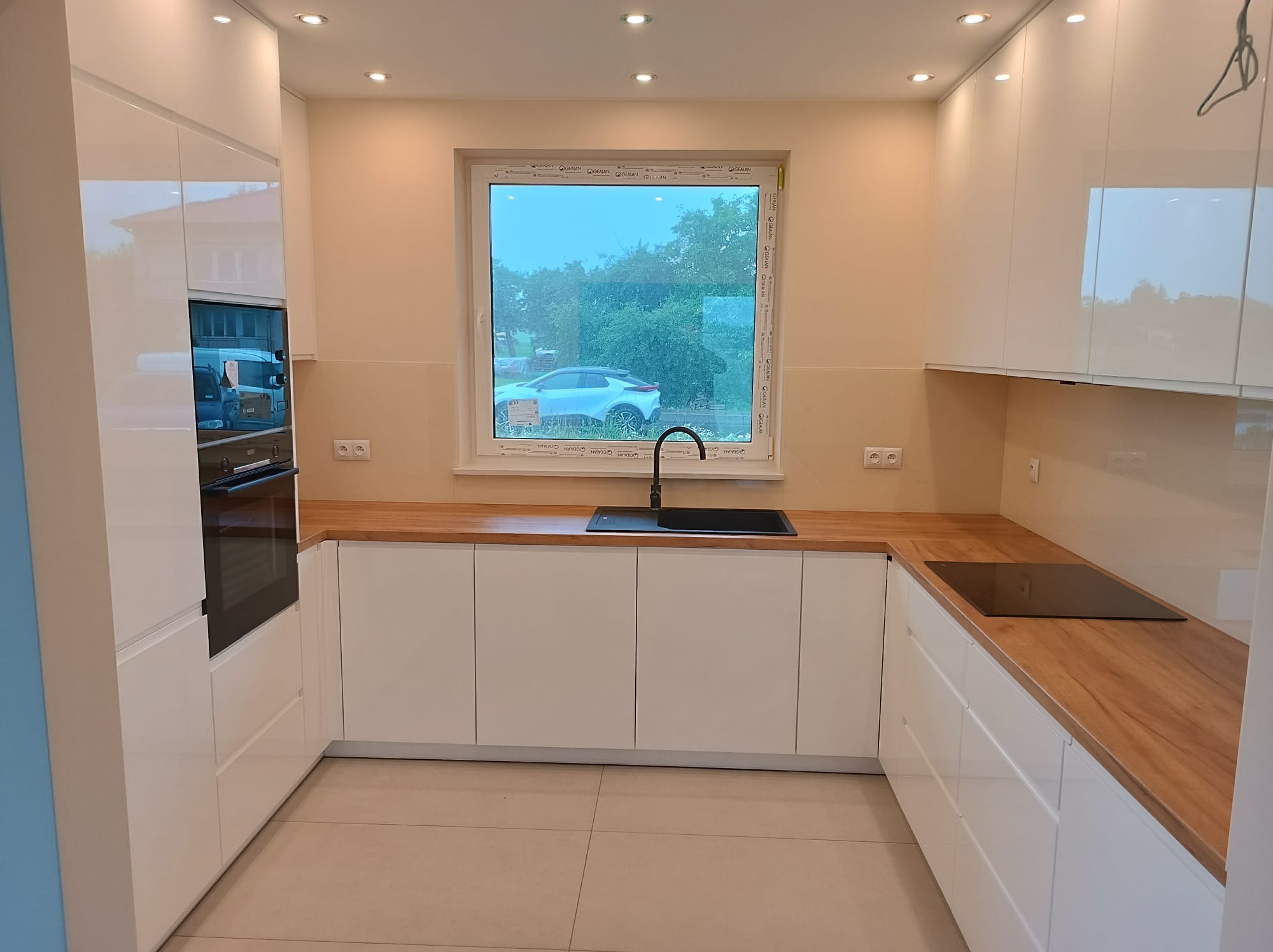 Biała, nowoczesna kuchnia z drewnianym blatem i oknem z widokiem na zieleń. Minimalistyczny design, jasne wnętrze, płyta indukcyjna i piekarnik w zabudowie.