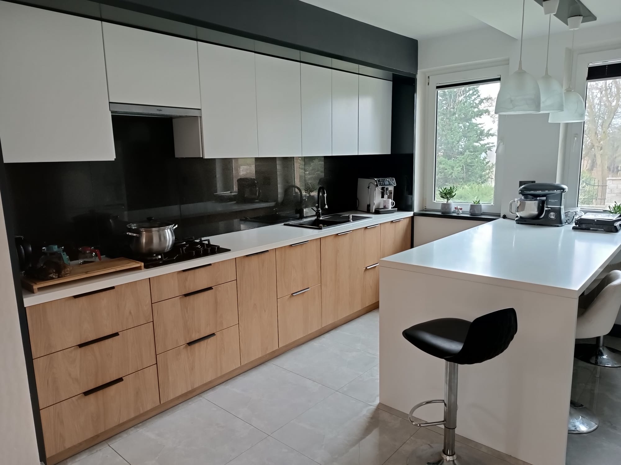 Nowoczesna kuchnia z białymi szafkami górnymi i drewnianymi dolnymi. Czarny panel nad blatem, wyspa z hokerem. Minimalistyczny design, jasne wnętrze.