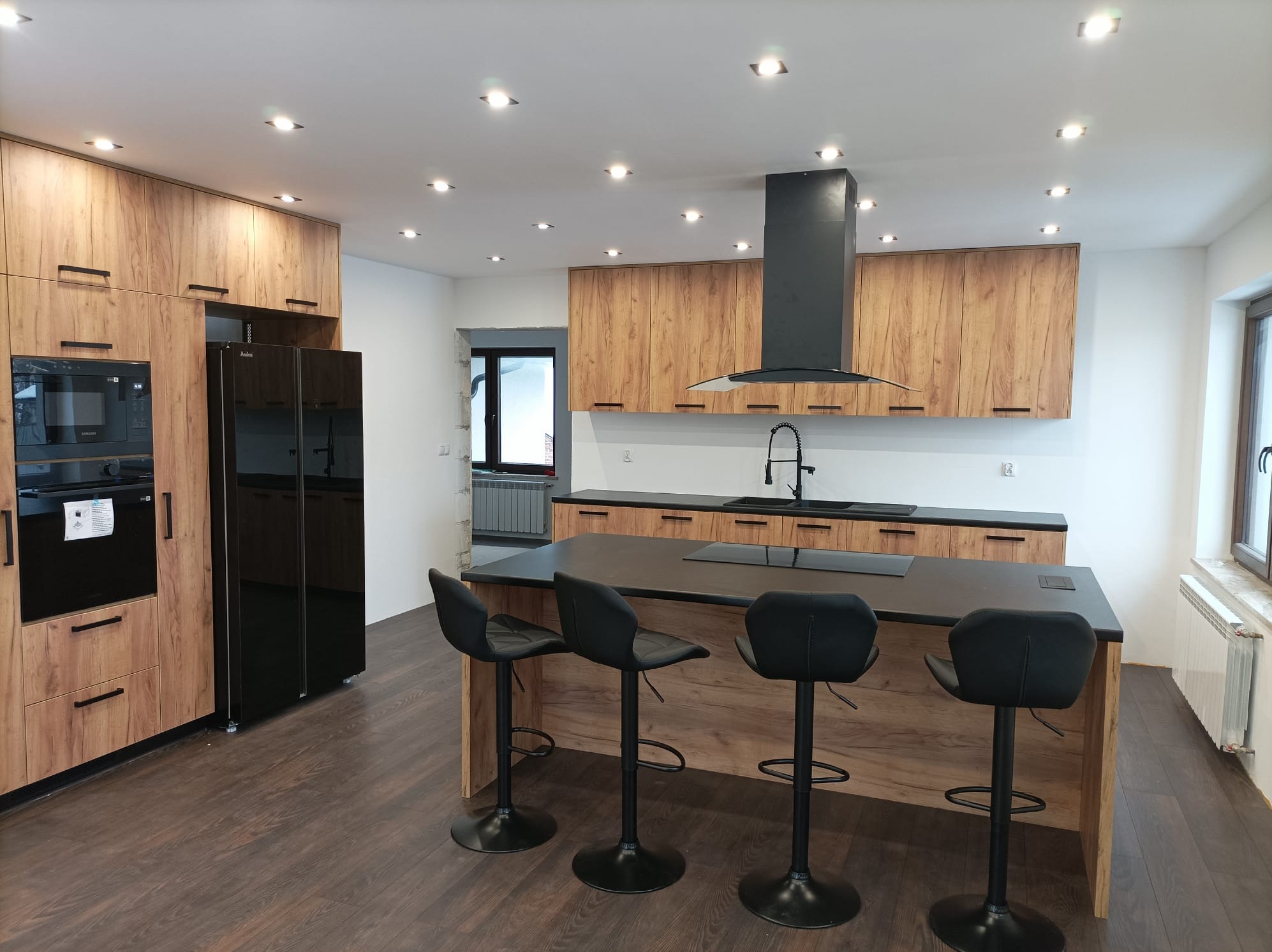 Nowoczesna kuchnia z drewnianymi szafkami, czarnym okapem i wyspą z czarnym blatem oraz trzema hokerami. Podłoga z paneli imitujących drewno. Oświetlenie punktowe.