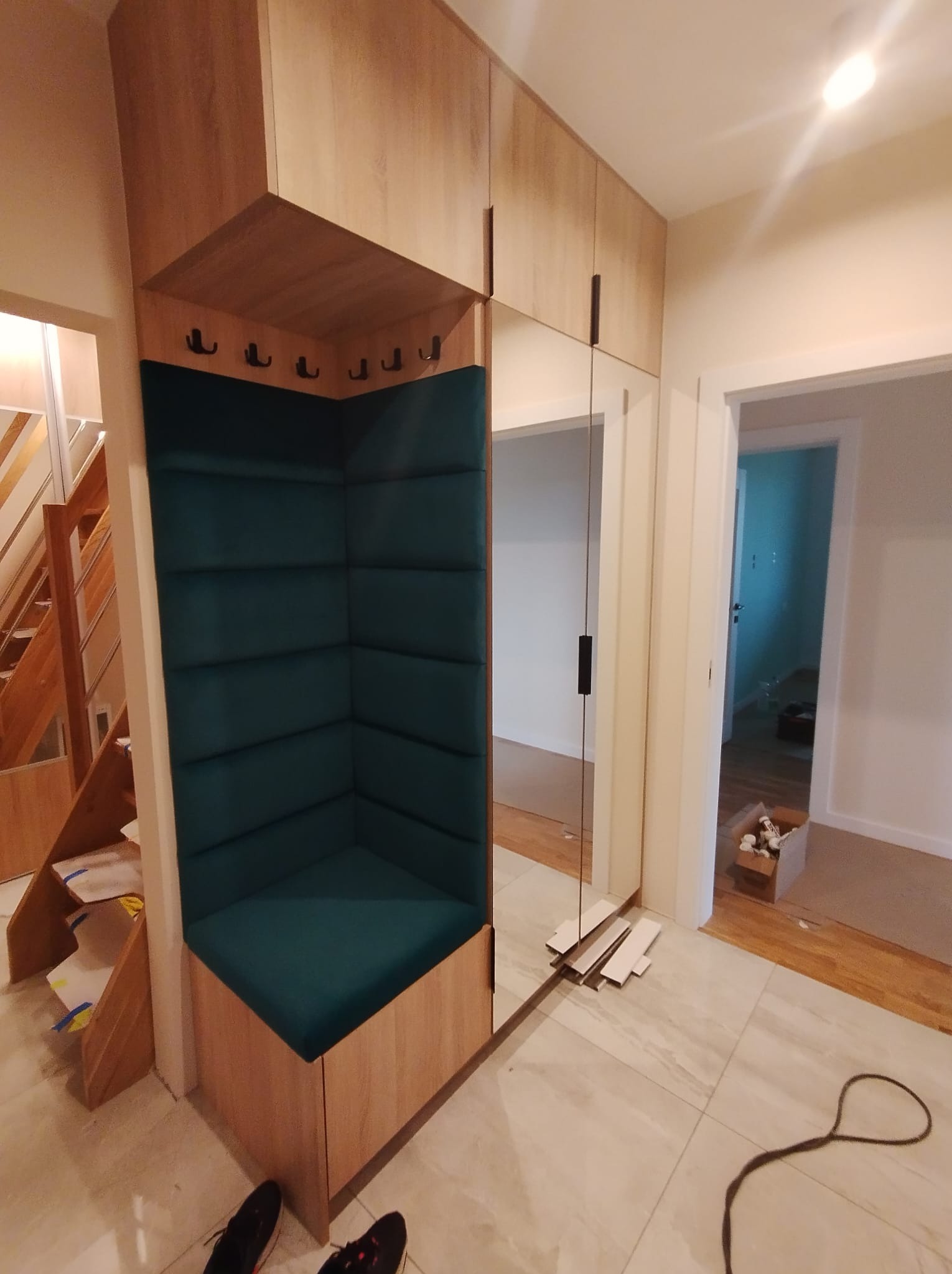 Funkcjonalna zabudowa przedpokoju z siedziskiem obitym butelkową zielenią, wbudowanym lustrem i szafkami z jasnego drewna. Widoczne schody i fragment podłogi.