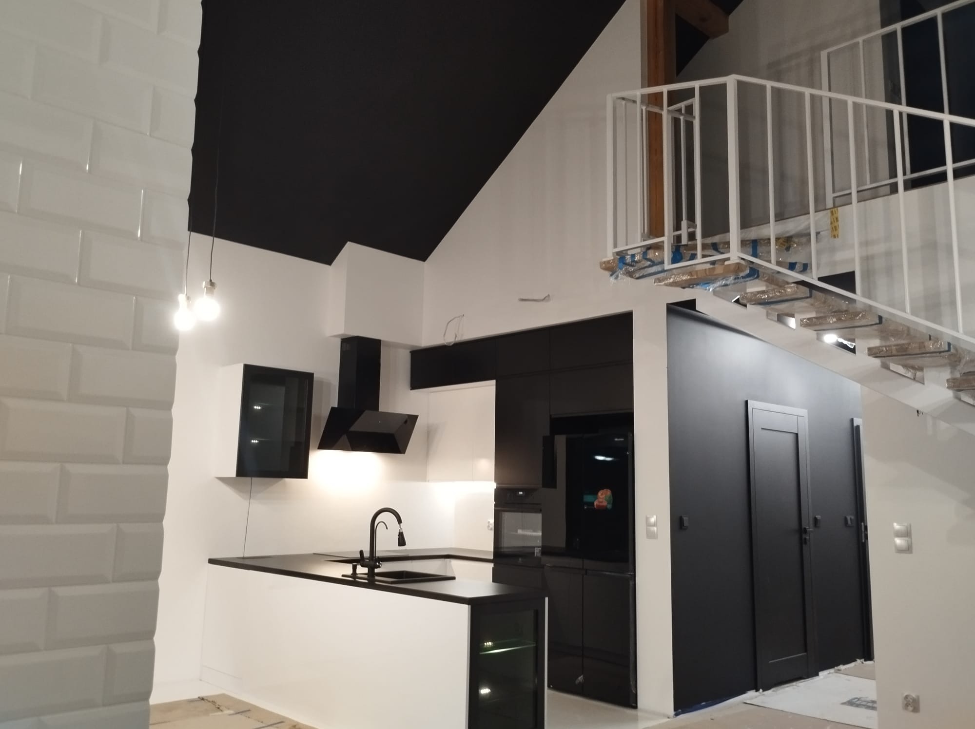 Nowoczesna kuchnia z czarnymi frontami i białymi elementami, oświetlona lampami wiszącymi, z widocznymi schodami z metalową balustradą w tle.
