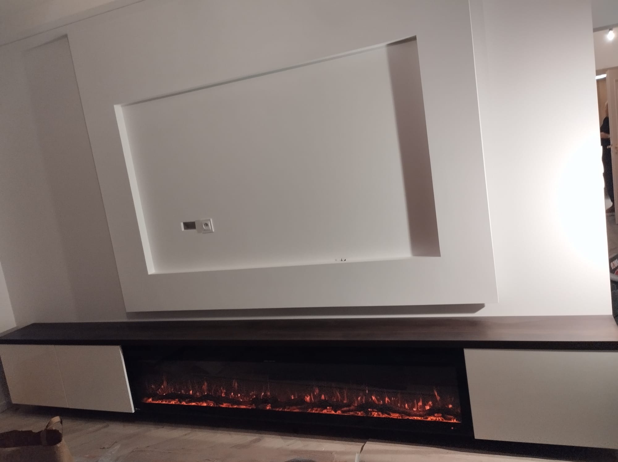 Nowoczesna zabudowa ściany z elektrycznym kominkiem i szafkami w kolorze ciemnego drewna i bieli. Minimalistyczny design z geometrycznym akcentem.