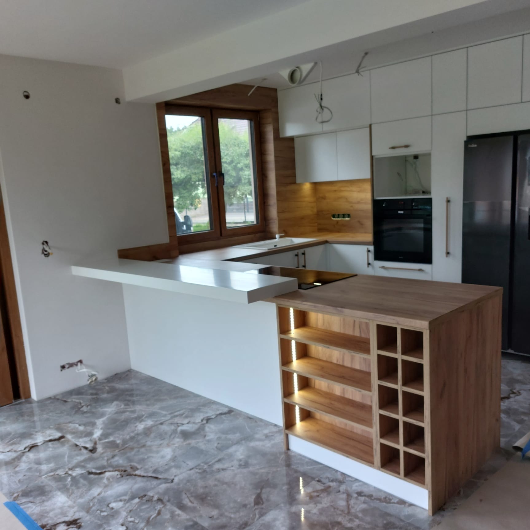 Nowoczesna kuchnia z wyspą i oświetleniem LED, drewniane akcenty, białe fronty, marmurowe płytki podłogowe. Widok na okno z zielenią. Częściowo widoczne instalacje.