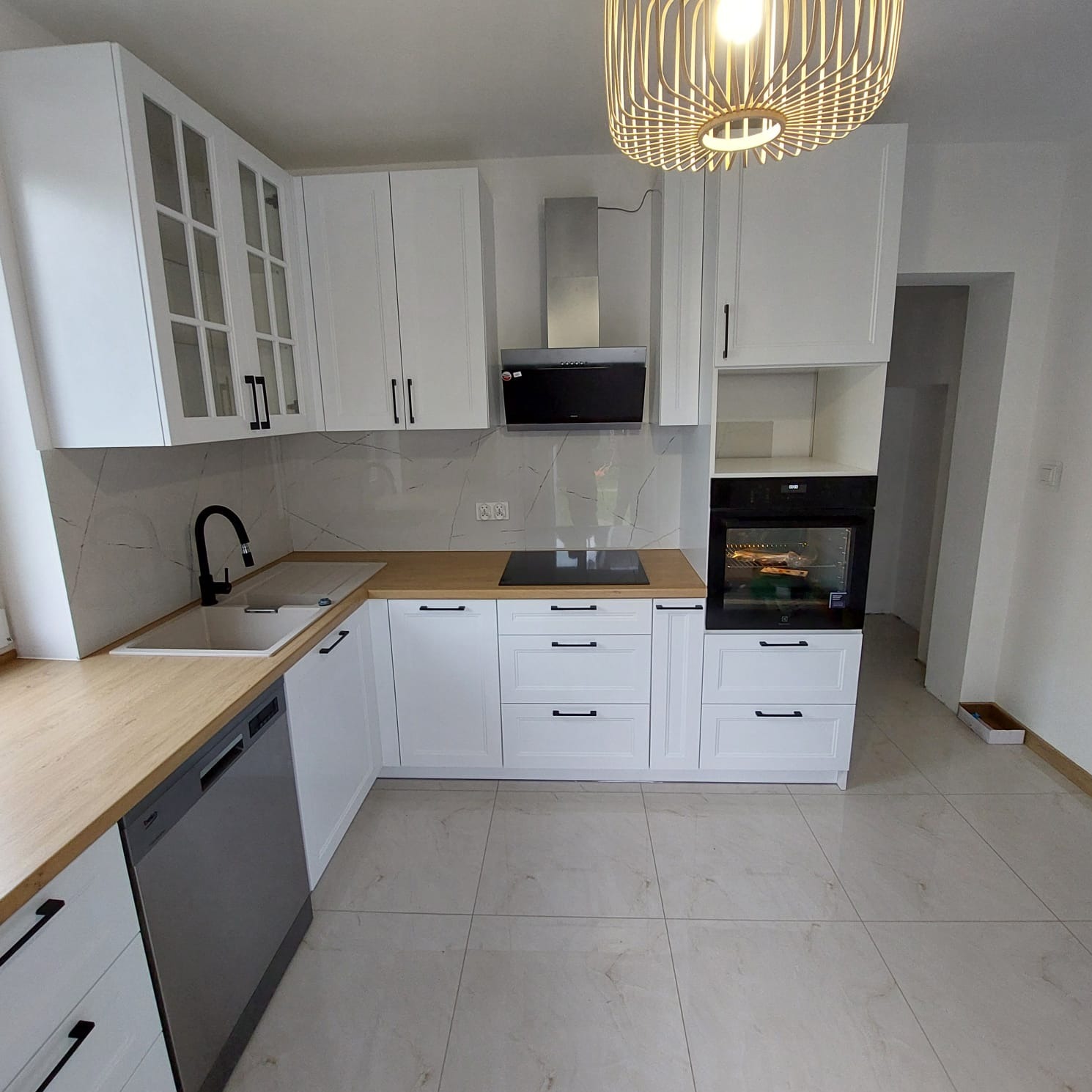 Biała kuchnia z drewnianym blatem i czarnymi uchwytami w Modliborzycach. Widoczna zabudowa kuchenna, piekarnik i płyta indukcyjna. Minimalistyczny design, jasne wnętrze.
