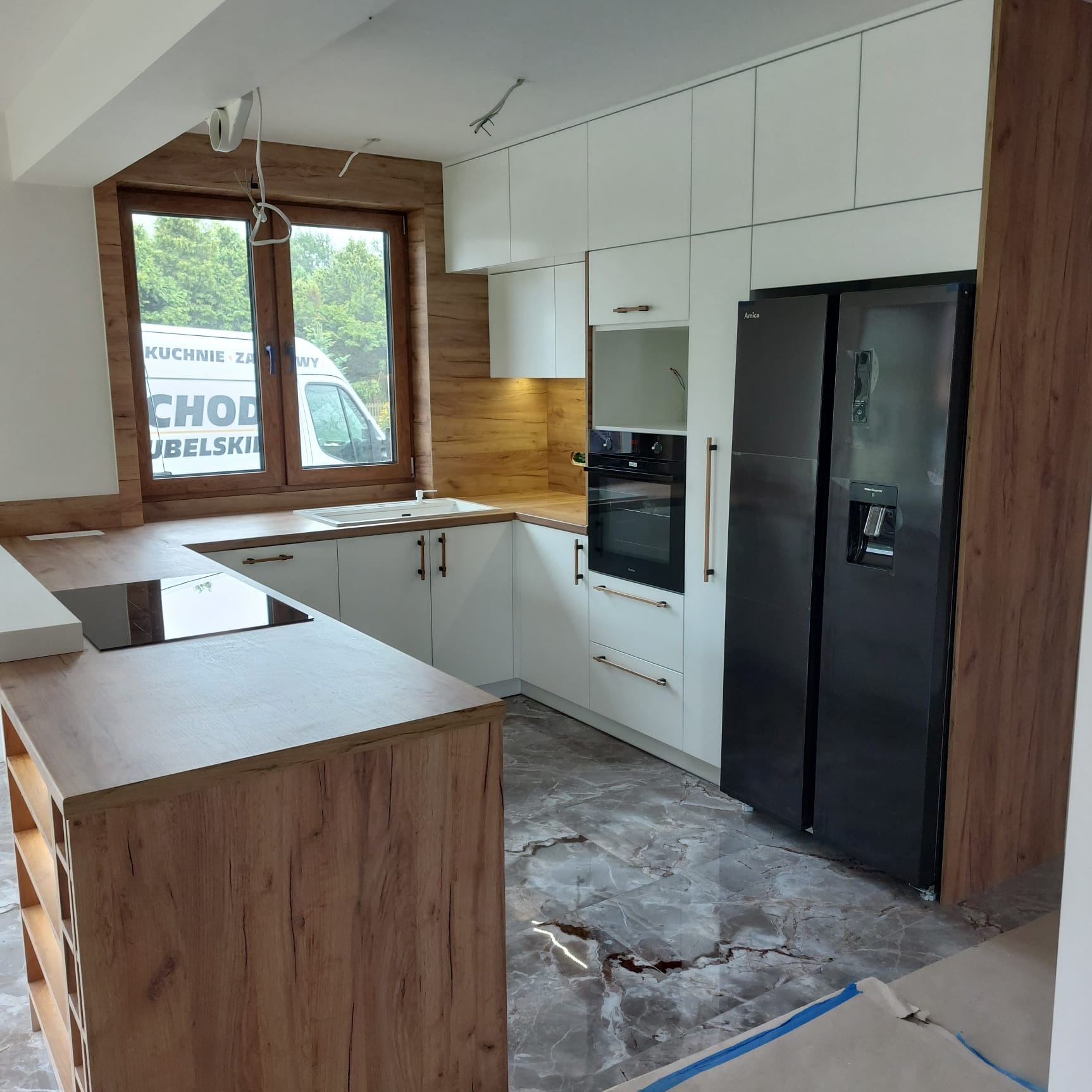 Nowoczesna kuchnia z białymi szafkami i drewnianymi akcentami, czarna lodówka Amica, marmurowe płytki podłogowe. Widok na okno z reklamą firmy.