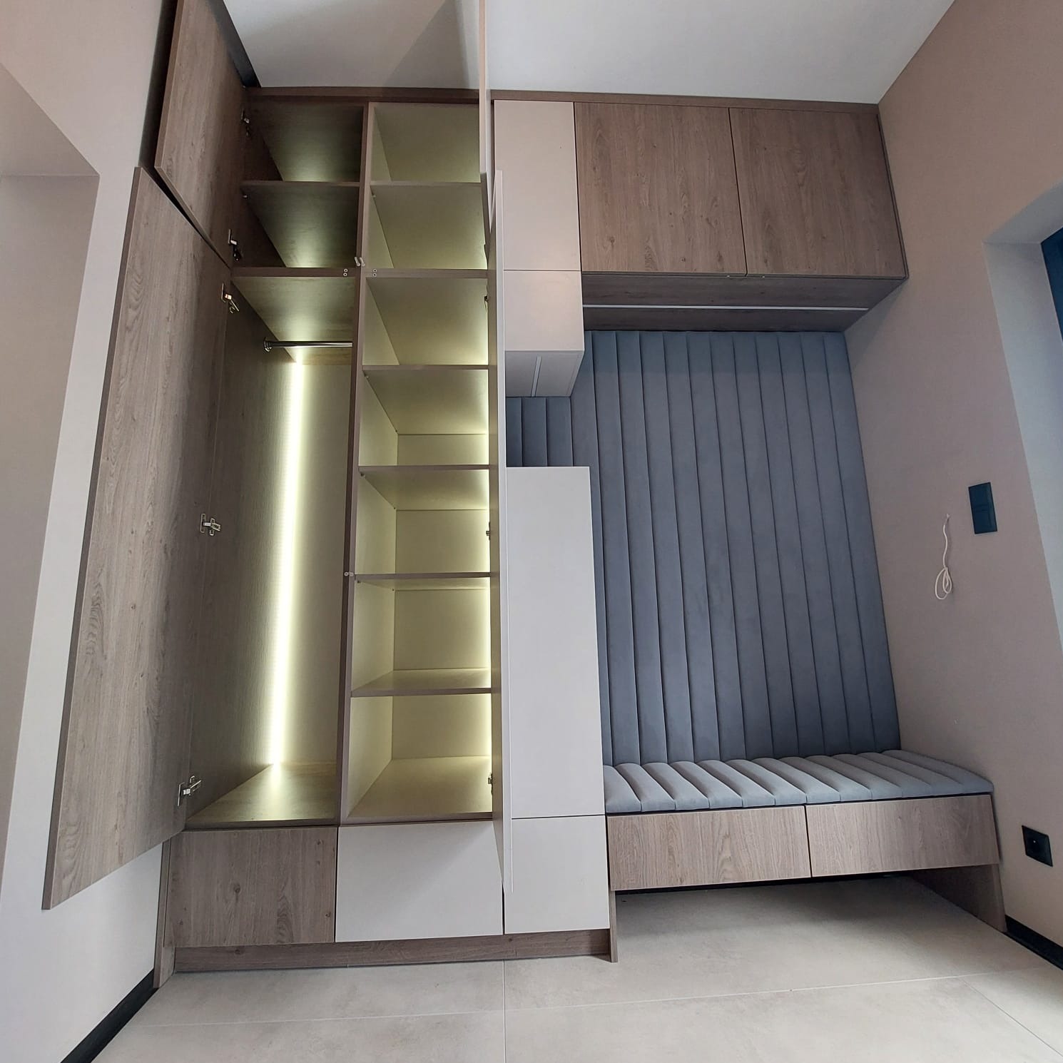 Funkcjonalna zabudowa wnęki: szafa z oświetleniem LED i tapicerowane siedzisko w odcieniach szarości i bieli, z elementami drewna. Nowoczesny design.