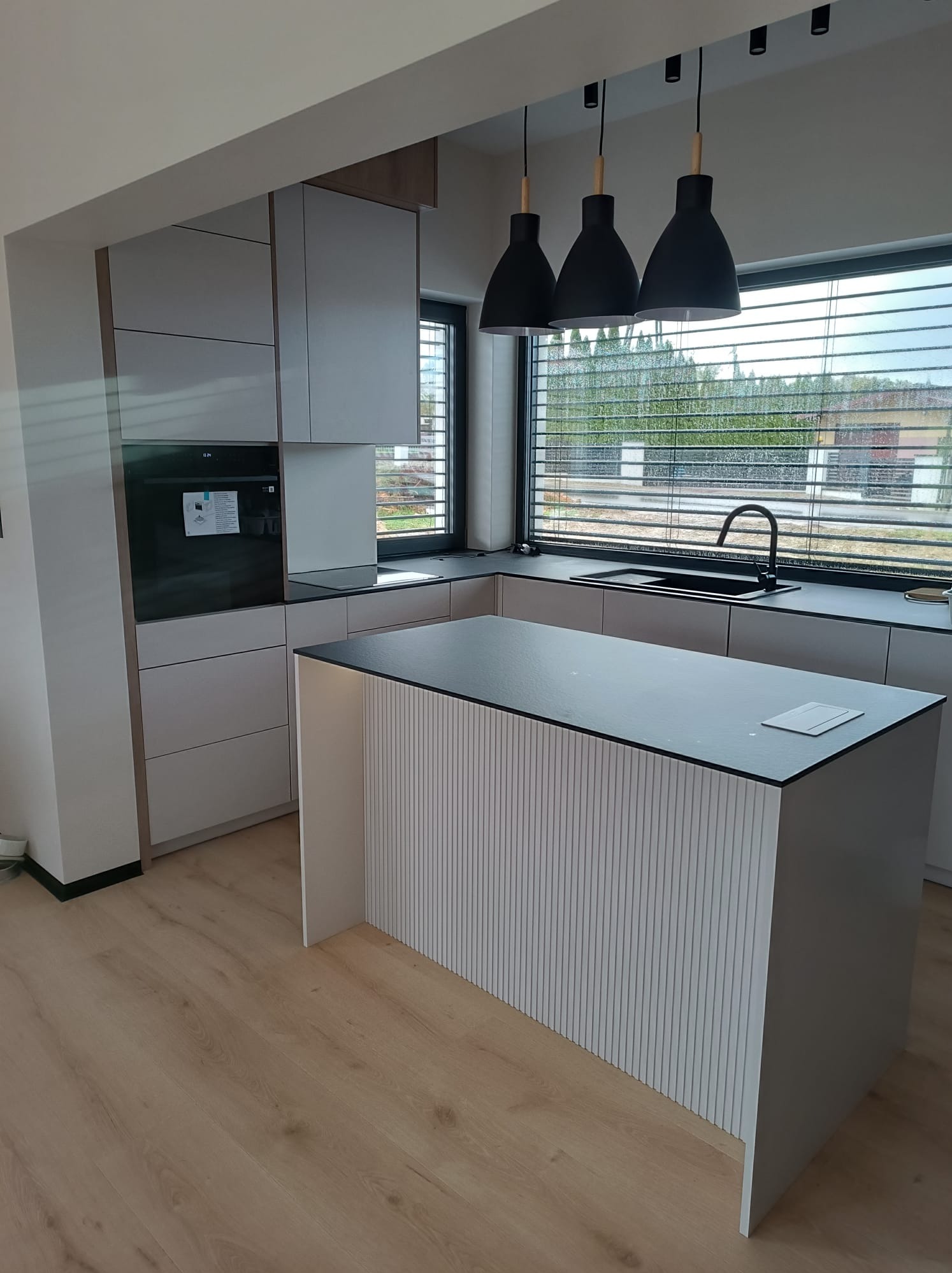 Nowoczesna kuchnia z wyspą: szare fronty, czarny blat, wiszące lampy. Minimalistyczny design, jasna podłoga, okno z żaluzjami. Detal: ryflowany front wyspy.