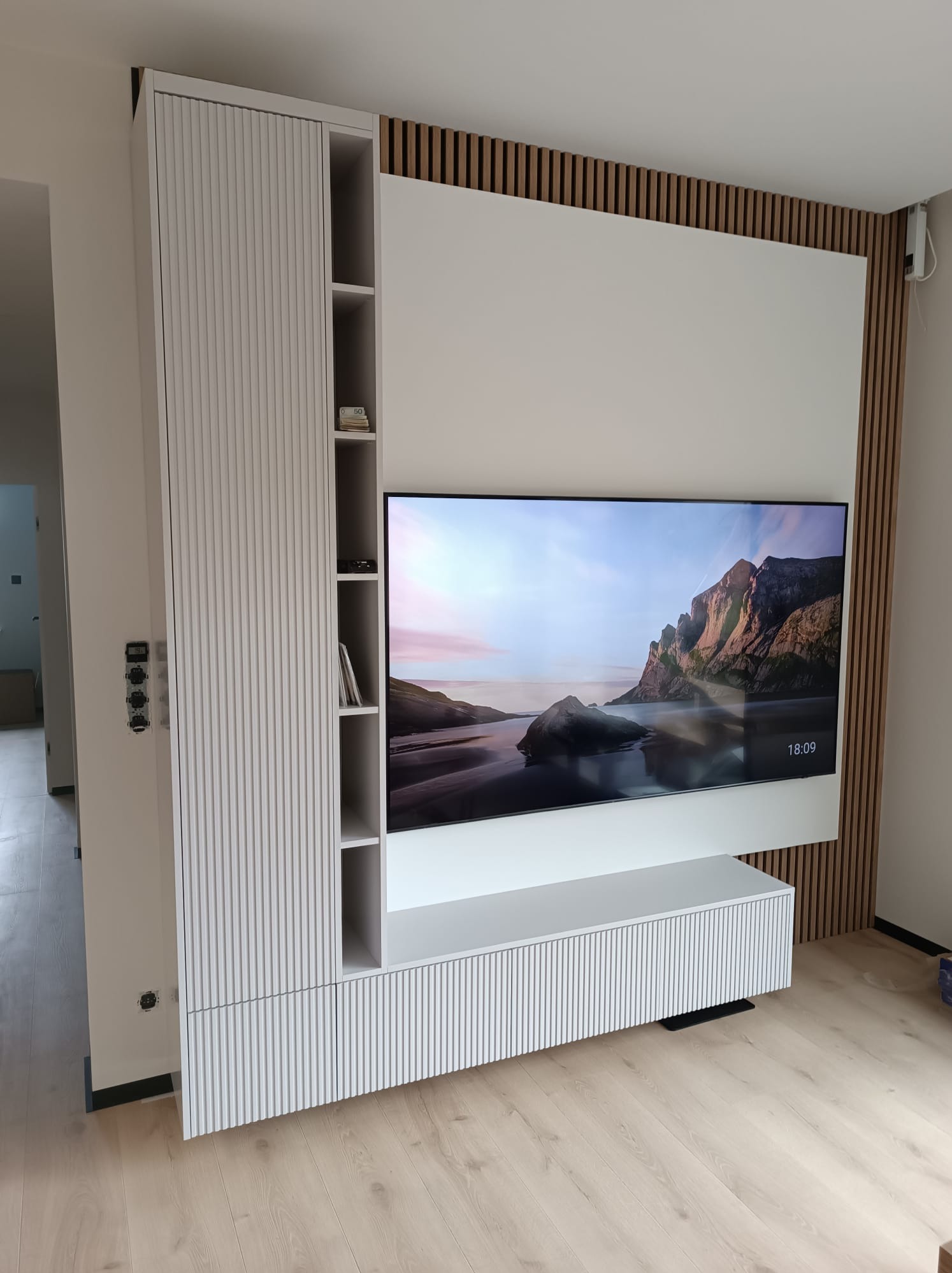 Biała zabudowa RTV z lamelami i szafką wiszącą, zintegrowana z drewnianą ścianką działową. Nowoczesny design, minimalistyczny styl, widok z perspektywy podłogi.