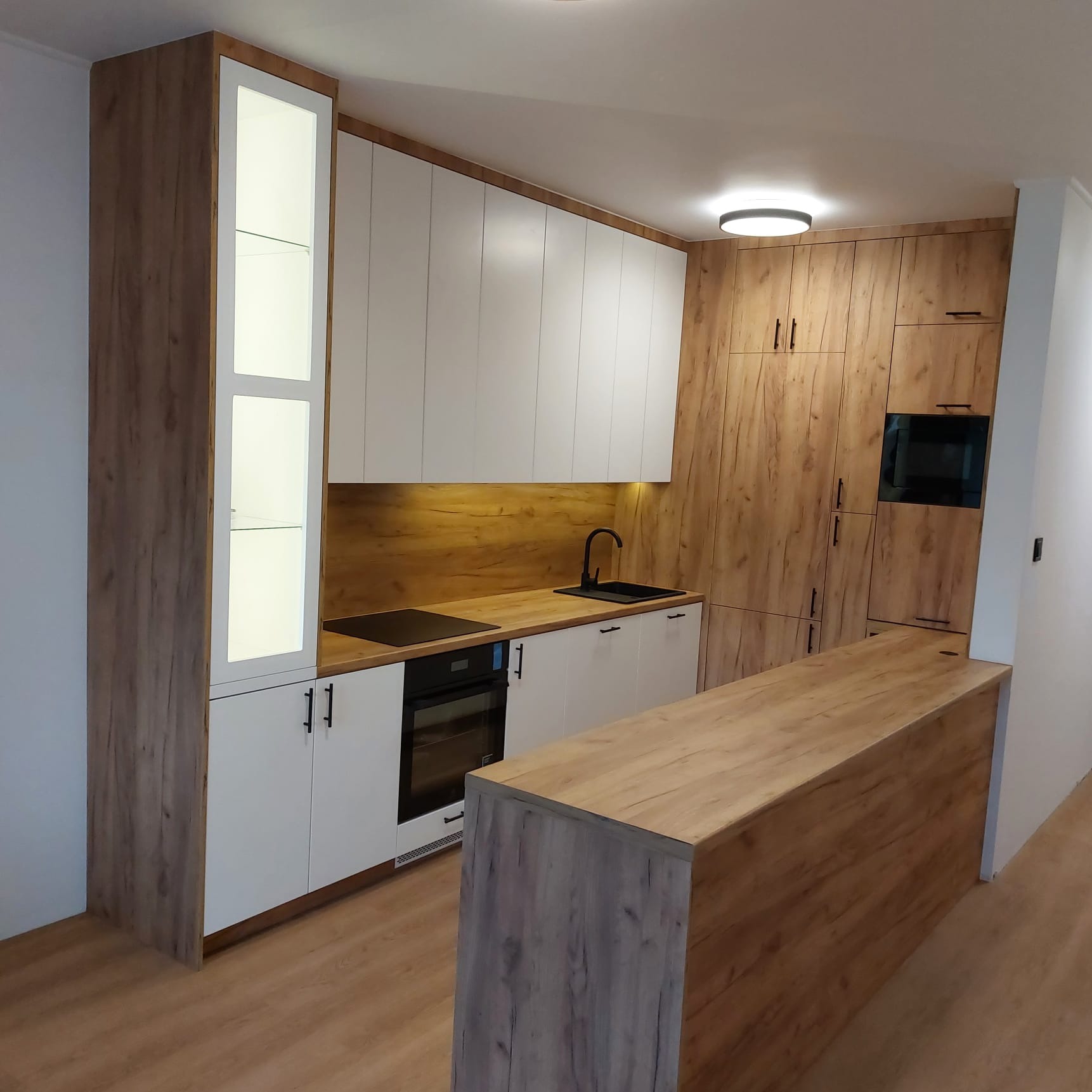Nowoczesna kuchnia z białymi frontami i drewnianymi akcentami. Wysoka zabudowa z oświetleniem LED, czarny zlew i płyta indukcyjna. Wyspa kuchenna z blatem w kolorze drewna.
