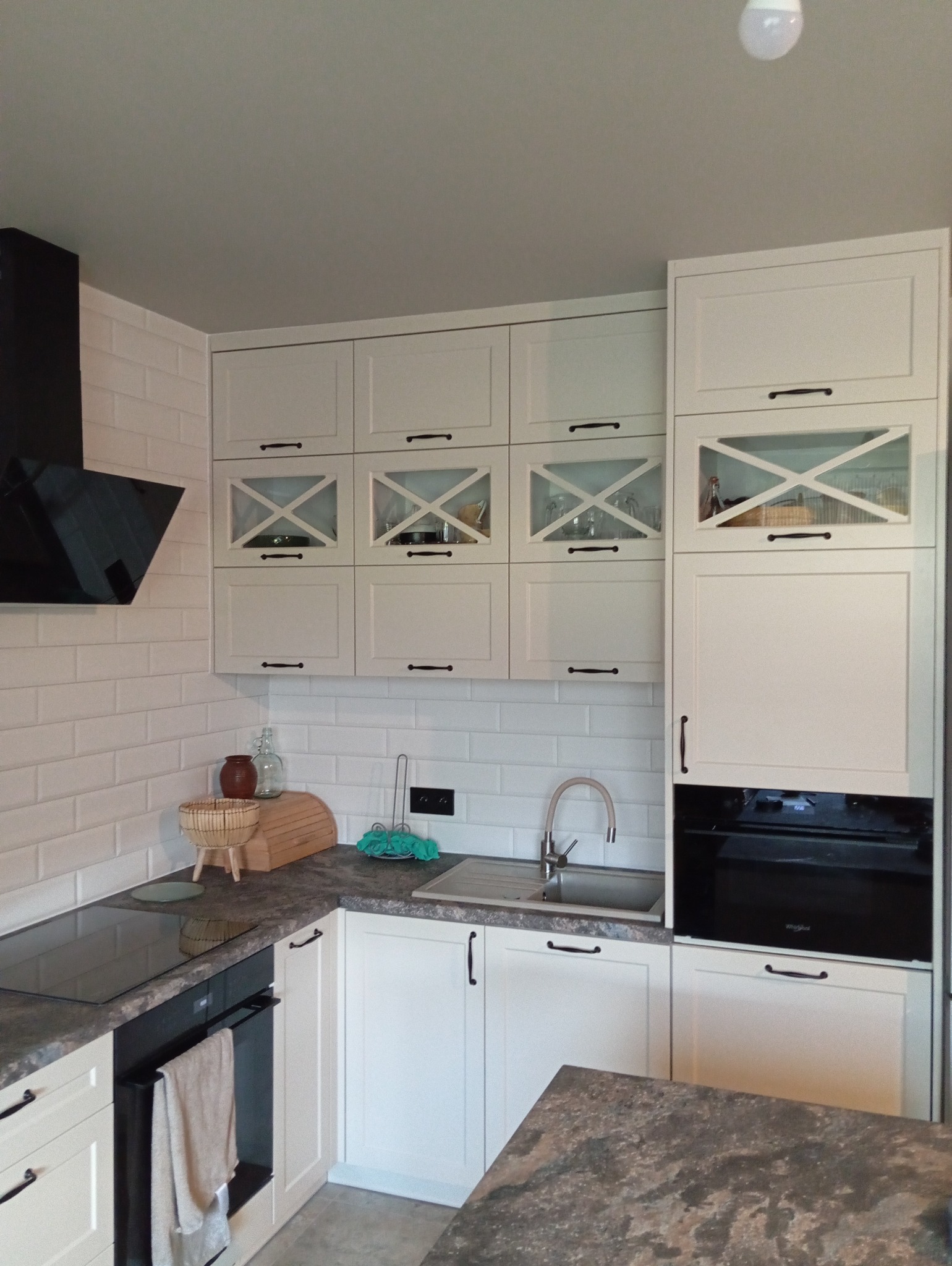 Jasna kuchnia z białymi szafkami, czarnymi uchwytami i szklanymi witrynami z krzyżowym wzorem. Płytki imitujące cegłę na ścianie i czarny okap.