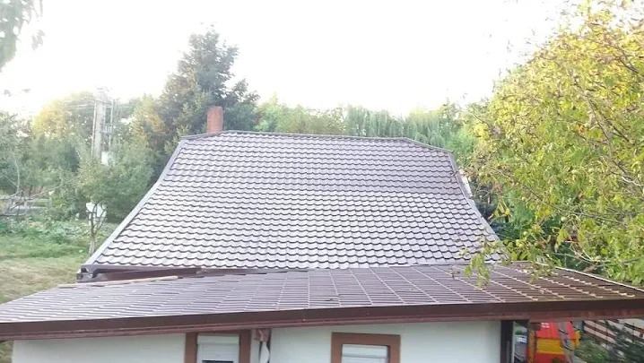 Brązowy dach z blachodachówki na tle zieleni. Widoczne rynny w kolorze dachu. Ujęcie z góry, lekko z prawej strony. Widoczny komin i drzewa w tle.