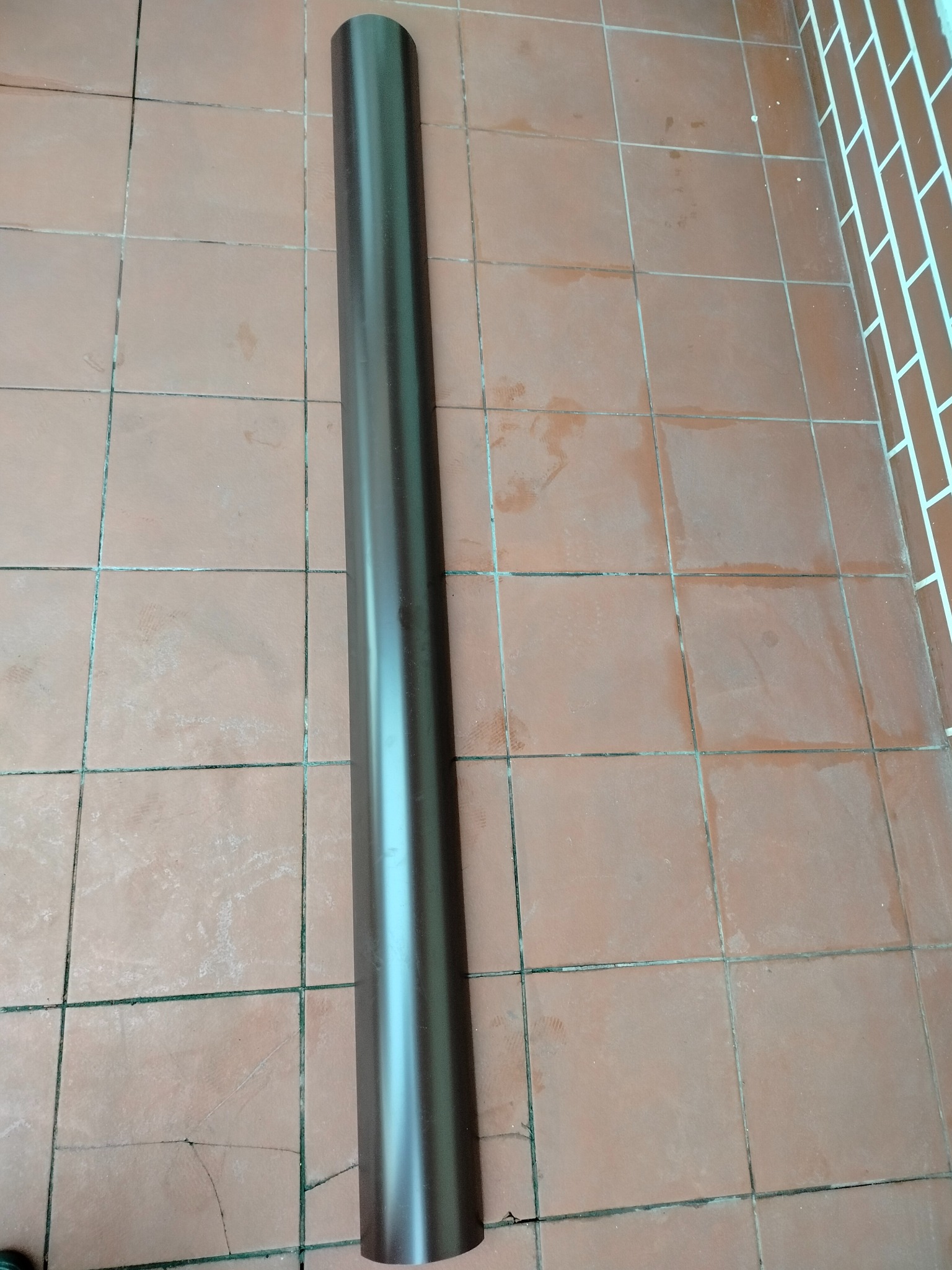 Brązowa rynna spustowa leży na terakocie w Krakowie. Element systemu odprowadzania wody deszczowej, przygotowany do montażu na dachu budynku.