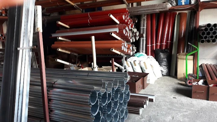 Magazyn z rynnami PCV w różnych kolorach i kształtach, ułożonymi na regałach. Widoczne także worki i kartony z akcesoriami.