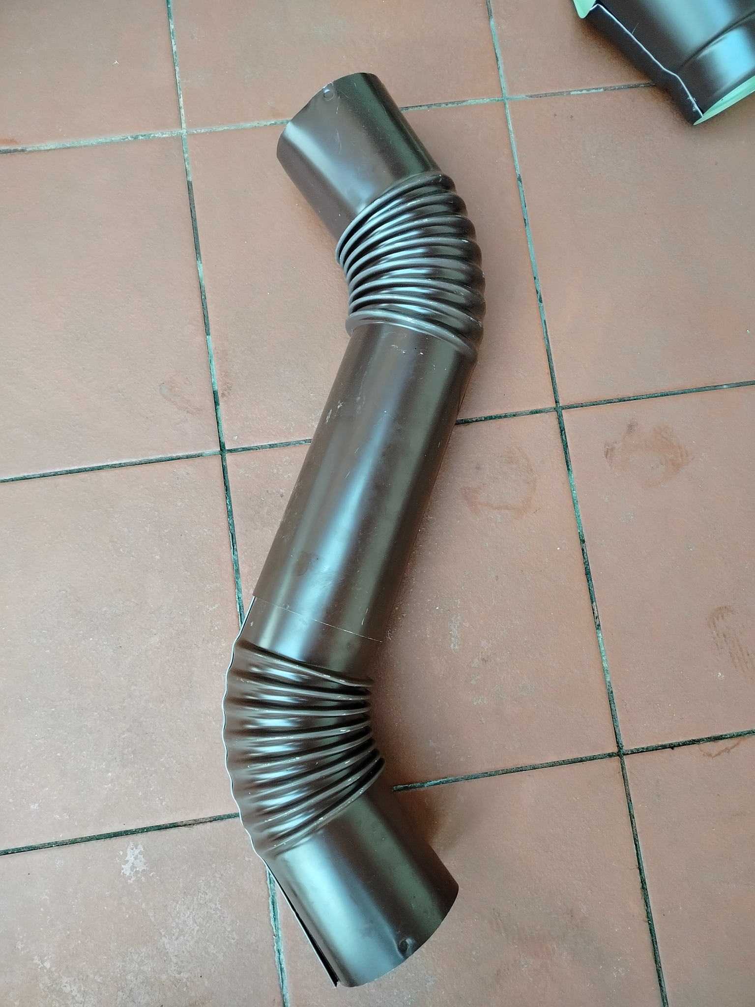 Brązowa, elastyczna rura spustowa rynny leży na brązowych płytkach podłogowych. Widoczny fragment innej rury w tle. Ujęcie z góry.