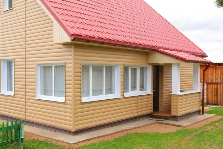 Dom z beżową elewacją i czerwonym dachem z blachy trapezowej. Widoczna rynna w kolorze dachu. Mały ganek z kolumnami. Ujęcie z poziomu trawnika.