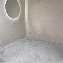 Remonty I Wykończenia Piotr Rak - Wykończone wnętrze: kremowe ściany z dekoracyjną fakturą, okrągłe lustro z podświetleniem, białe marmurowe płytki podłogowe i gniazdka elektryczne.