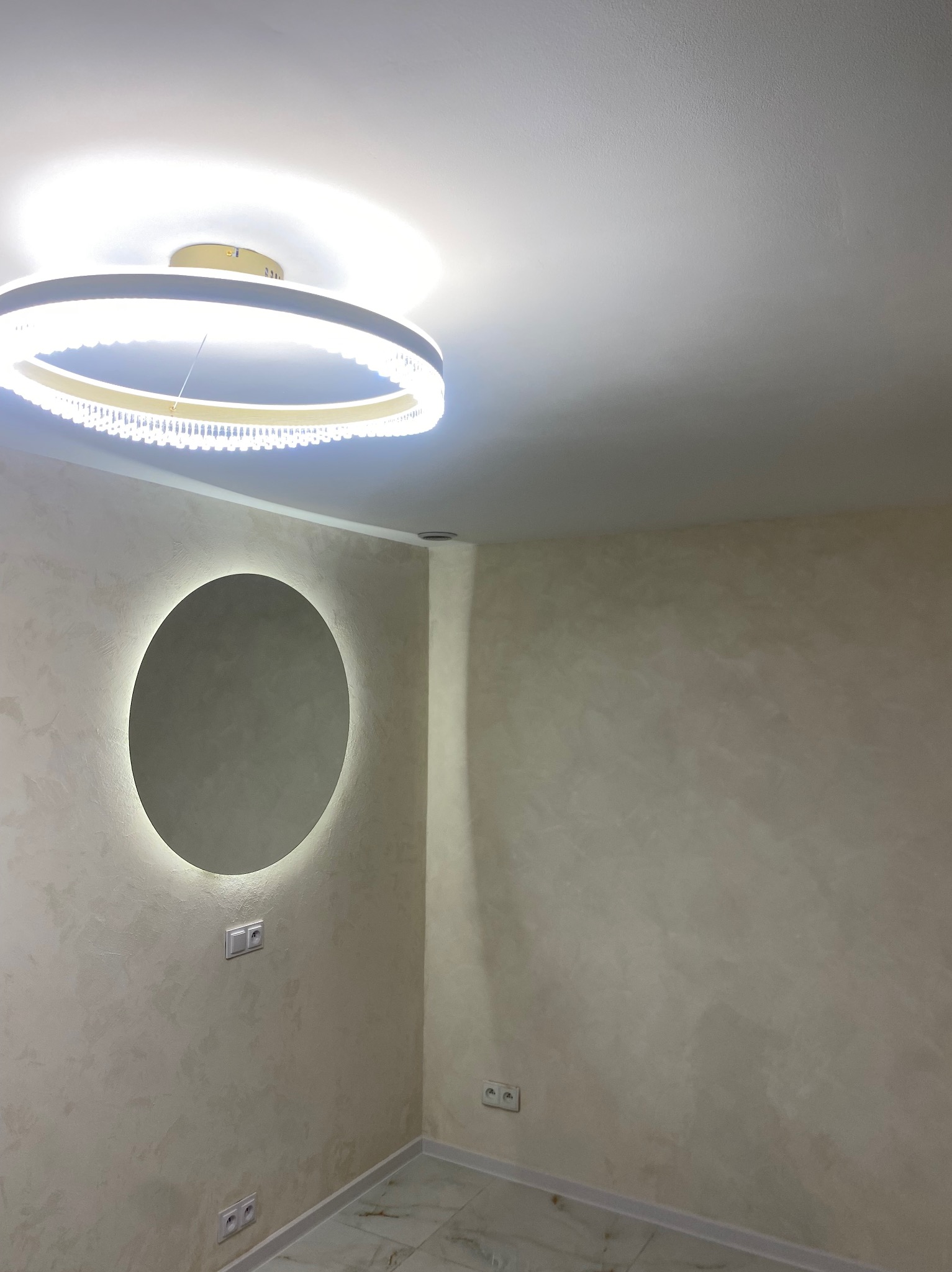 Wykończone wnętrze: ściany z dekoracyjną fakturą w odcieniach beżu, okrągłe lustro z podświetleniem LED i nowoczesna lampa sufitowa. Widoczne gniazdka elektryczne.