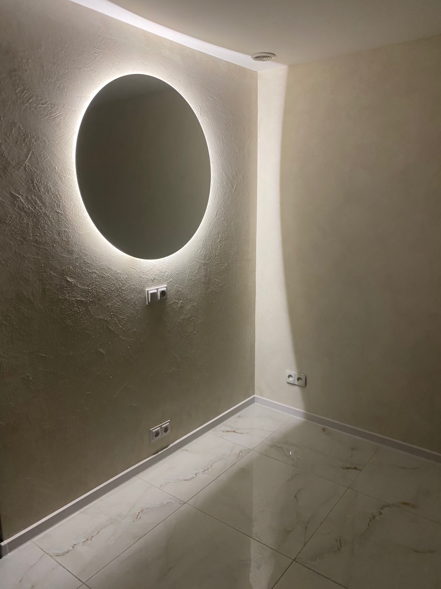 Wykończone wnętrze: kremowe ściany z fakturą, okrągłe lustro z podświetleniem LED, białe płytki podłogowe z marmurkowym wzorem i gniazdka elektryczne.