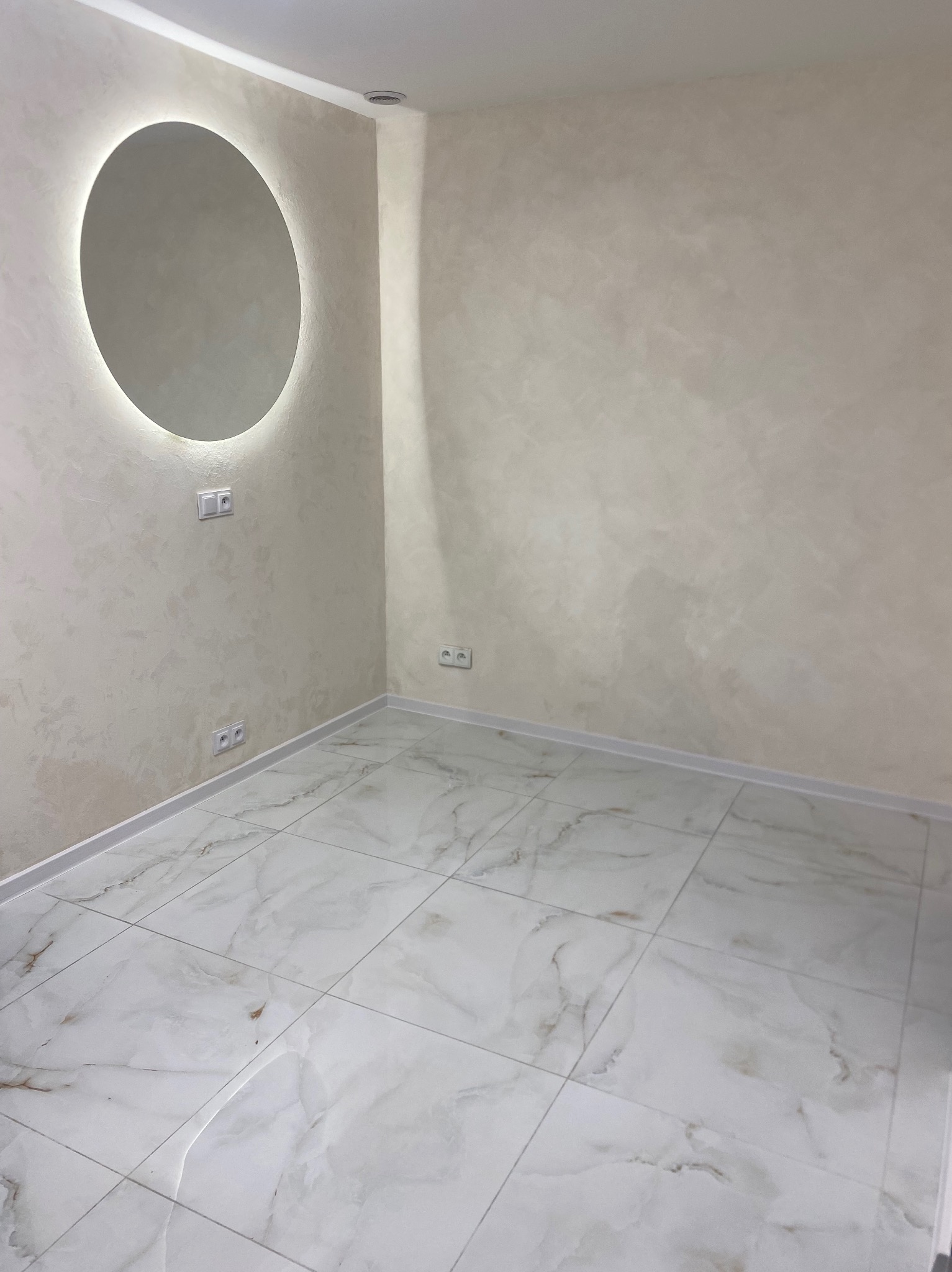 Wykończone wnętrze: kremowe ściany z dekoracyjną fakturą, okrągłe lustro z podświetleniem, białe marmurowe płytki podłogowe i gniazdka elektryczne.