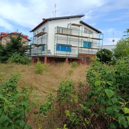 Teren Zieleni Vasyl Dutchak - Dom w trakcie ocieplania, otoczony rusztowaniem. Okna zabezpieczone folią. Na pierwszym planie zarośnięty ogród z wysoką trawą i krzewami.