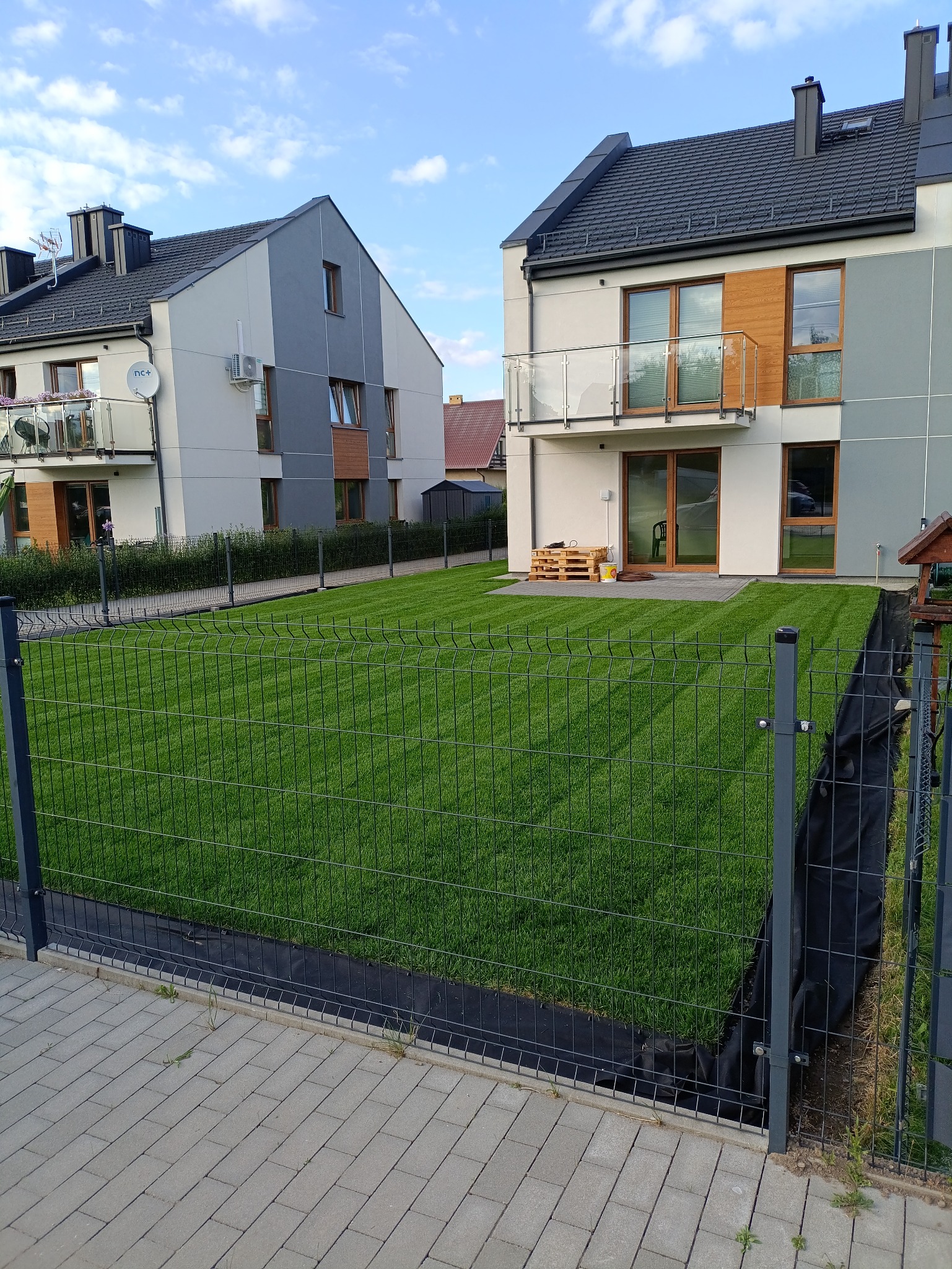 Świeżo skoszony trawnik przed nowoczesnym domem z szarą elewacją, ogrodzony metalową siatką. Widoczne regularne pasy po koszeniu i fragmenty kostki brukowej.