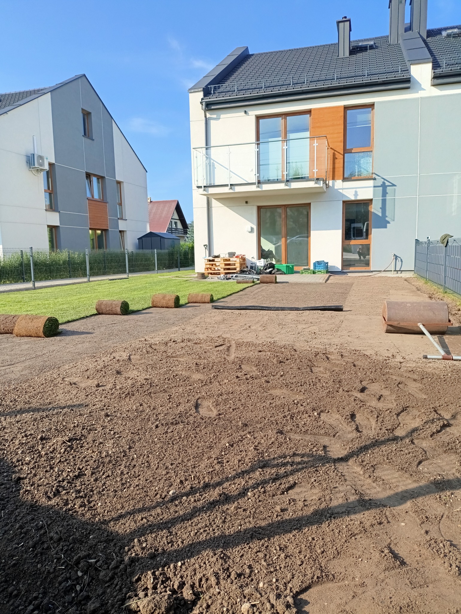 Rozplantowana ziemia pod trawnik z rolki, częściowo ułożona trawa w rolkach, walec do trawy i nowoczesne budynki w tle, słoneczny dzień.