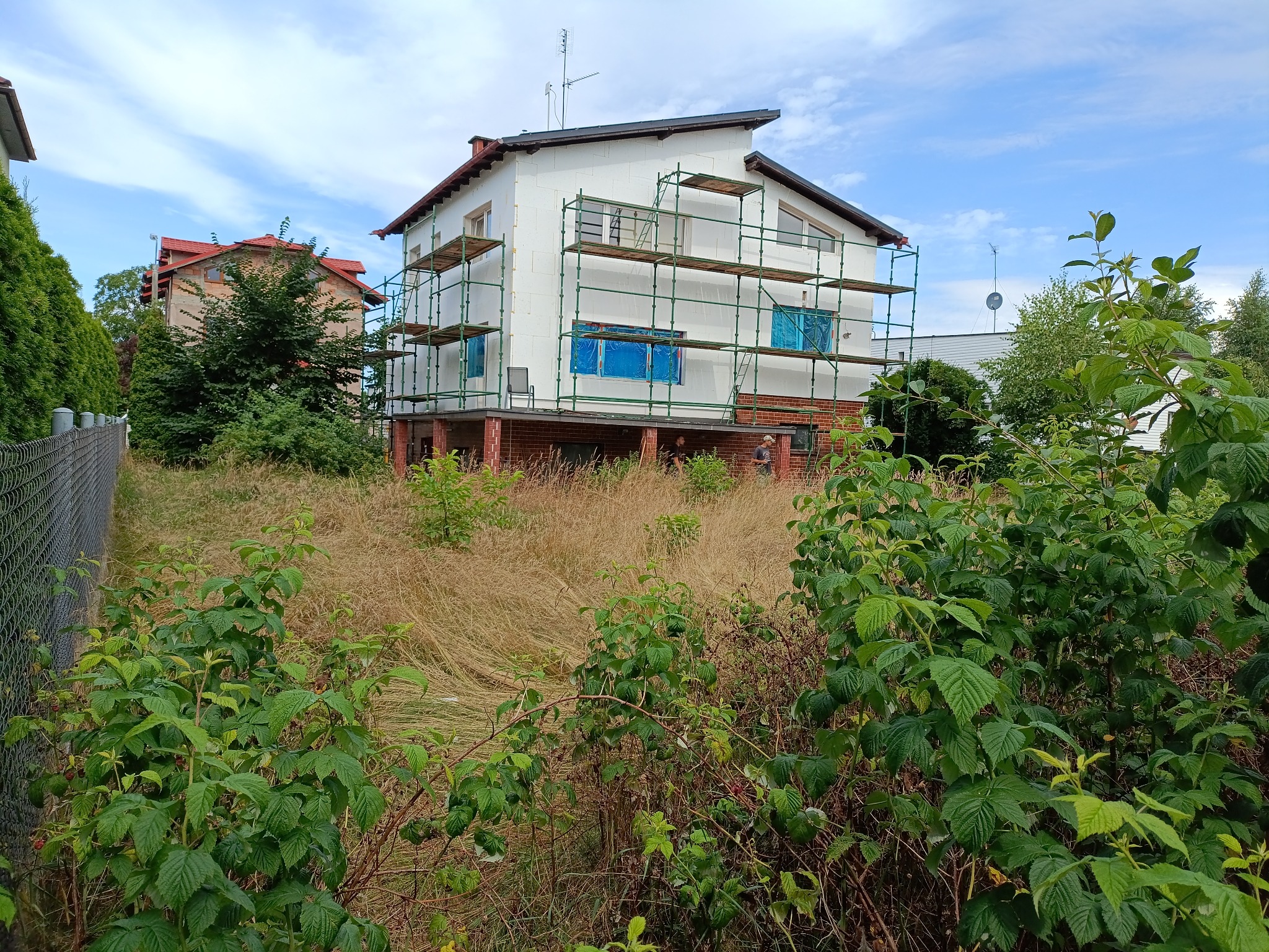 Dom w trakcie ocieplania, otoczony rusztowaniem. Okna zabezpieczone folią. Na pierwszym planie zarośnięty ogród z wysoką trawą i krzewami.