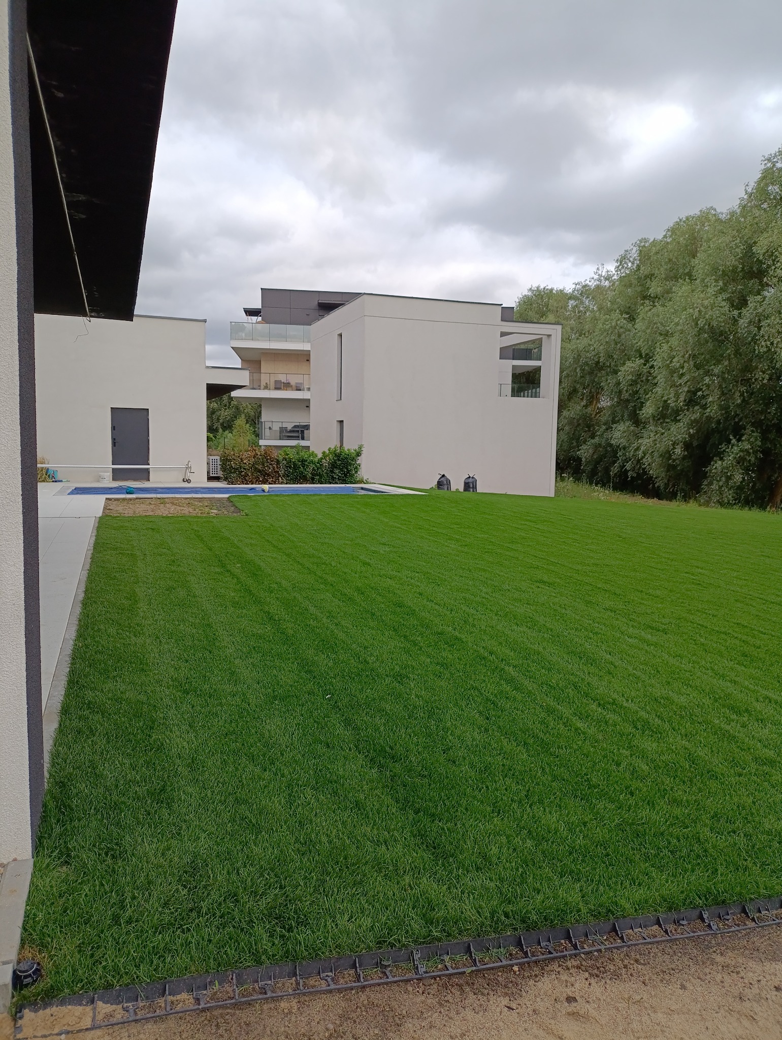 Perfekcyjnie skoszony trawnik przed nowoczesnym domem z basenem, widok z poziomu gruntu. Trawa równo przycięta, tworzy wzór w paski. Dwa worki stoją obok basenu.