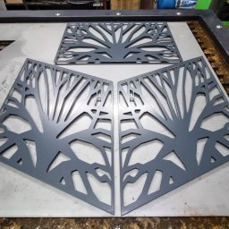 Krzysztof Purol welderone - Metalowe panele dekoracyjne z motywem drzewa, wycięte laserowo, leżące na metalowym stole warsztatowym. Dwa panele ustawione pod kątem, jeden u góry. Szary kolor.