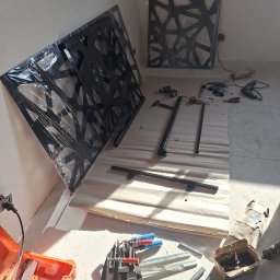 Krzysztof Purol welderone - Elementy metalowe z ażurowym wzorem, częściowo w folii, leżą na drewnianej powierzchni. Widoczne narzędzia i akcesoria montażowe, sugerujące etap przygotowania do instalacji.