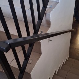 Krzysztof Purol welderone - Czarne, metalowe poręcze schodowe z pionowymi szczeblami, zamocowane do ściany i stopni. Widoczne ślady montażu, płytki schodowe w odcieniach brązu.