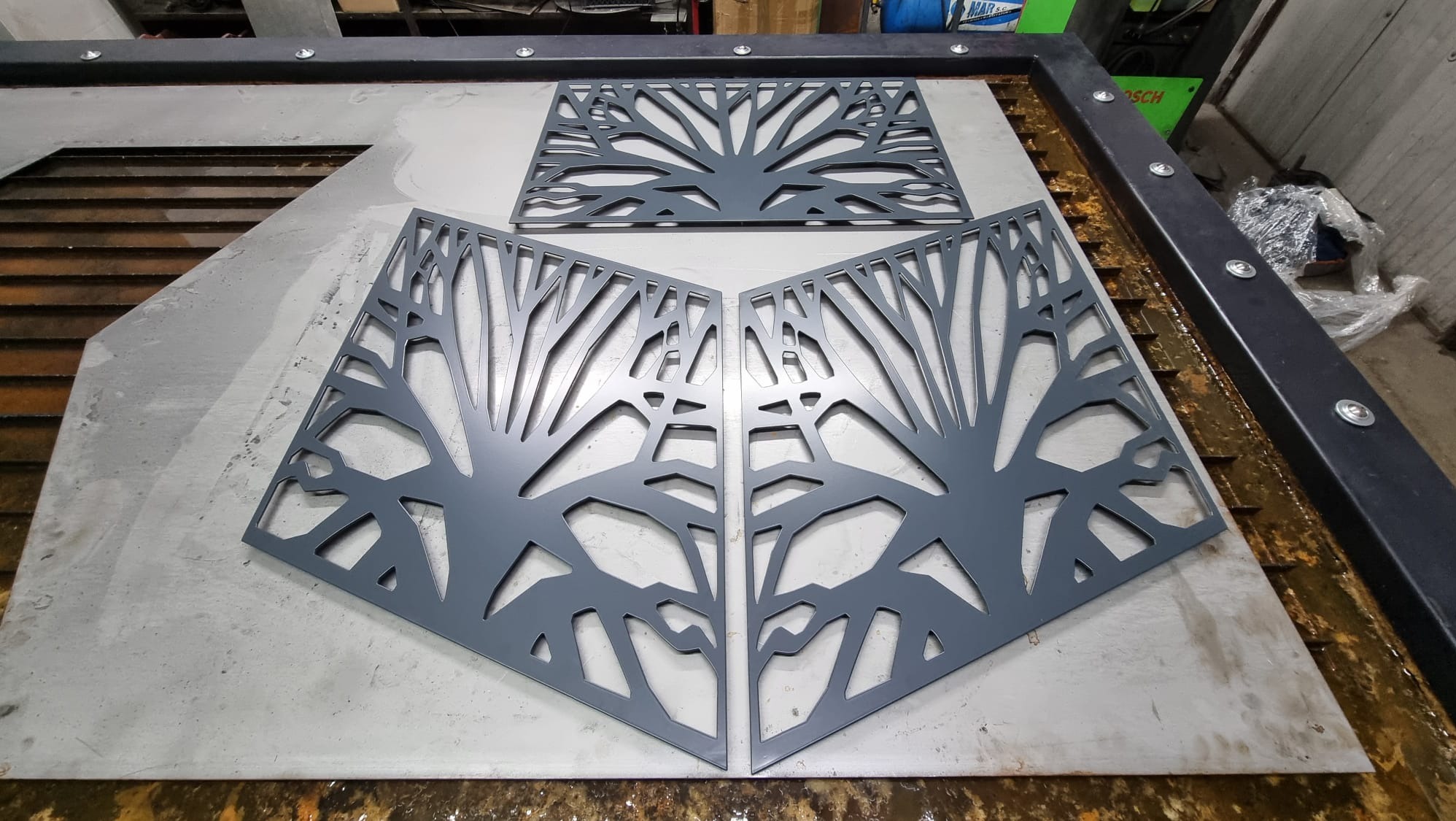 Metalowe panele dekoracyjne z motywem drzewa, wycięte laserowo, leżące na metalowym stole warsztatowym. Dwa panele ustawione pod kątem, jeden u góry. Szary kolor.