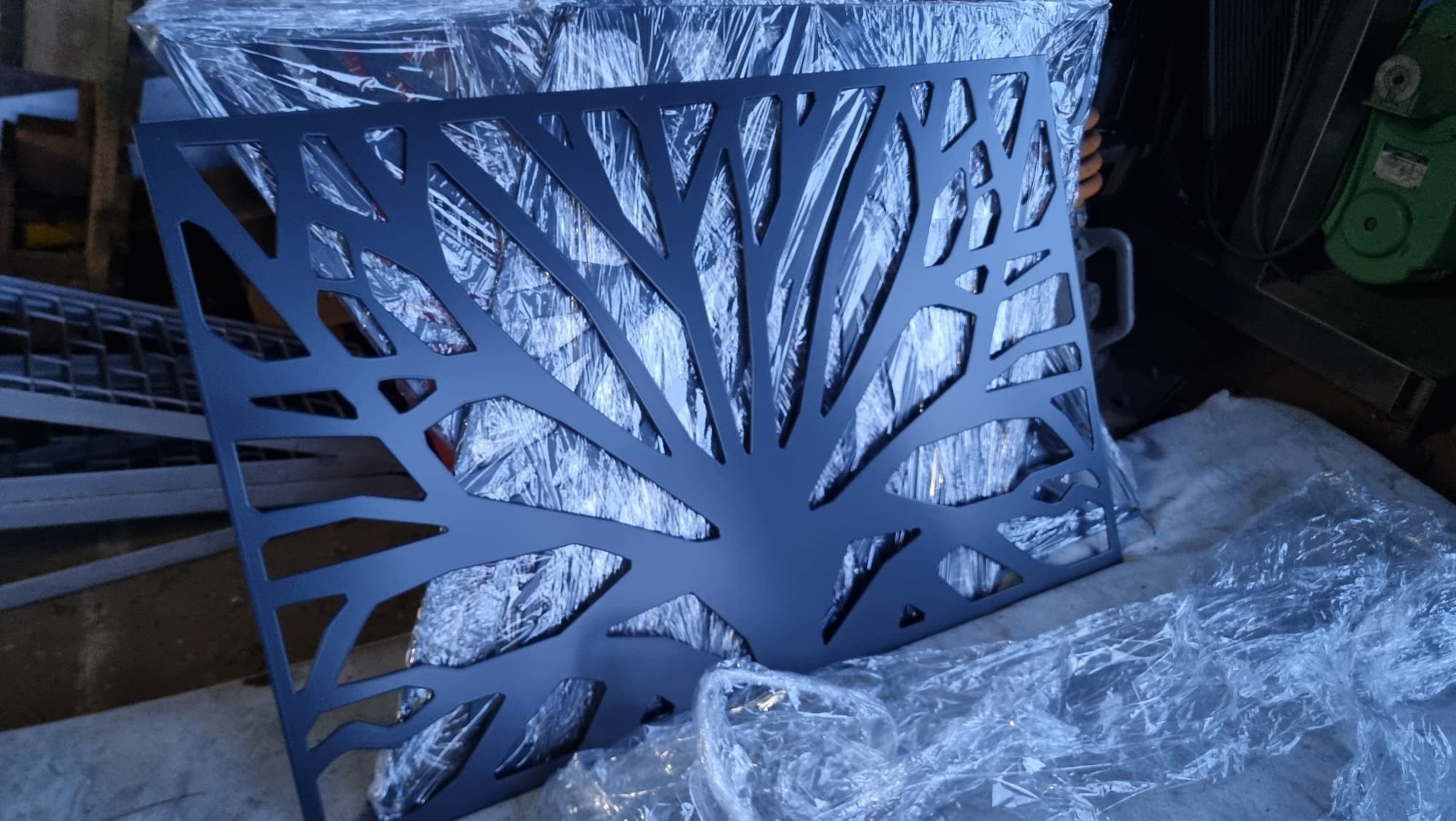 Metalowy panel dekoracyjny z wyciętym motywem drzewa, leżący na folii ochronnej. Ciemny kolor, precyzyjne wycięcia, widoczne elementy warsztatu w tle.